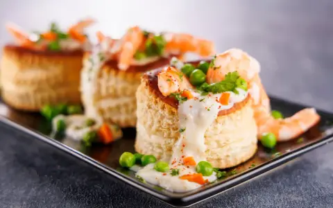 Trois bouchées de tapas crevette, garnies de crevettes, petits pois et sauce crémeuse, sur un plateau noir.