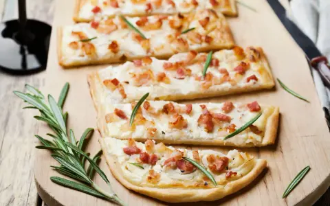 Quatre tartes flambées, une délicieuse entrée alsacienne, garnies de lardons et de romarin sur une planche en bois.