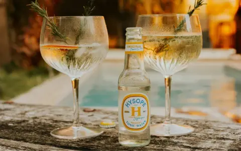 Une bouteille de tonic Hysopé Original, prête à être versée dans deux verres de gin rafraîchissants, décorés de romarin et d'une tranche d'orange.