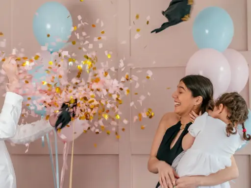 Explosion de confettis roses et dorés lors d'une fête pour annoncer fille ou garçon. Une femme rit, tenant un enfant, entourée de ballons bleus et roses.