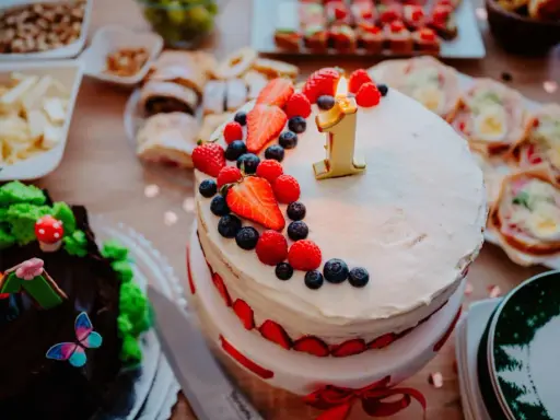 Gâteau à deux étages décoré de fruits rouges et d'une bougie "1" pour un déjeuner d'anniversaire. Un autre gâteau chocolaté, des petits fours et des fruits sont disposés sur la table.