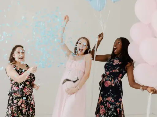 Trois femmes célèbrent un gender reveal, avec des confettis bleus et des ballons roses et bleus. C'est quoi ce moment joyeux !