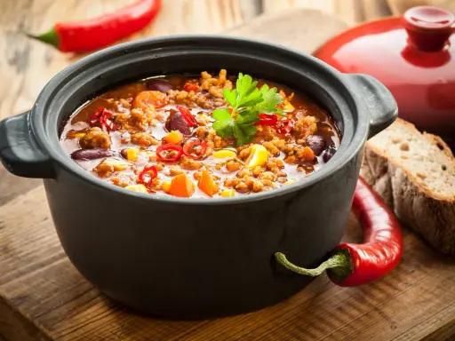 Un plat de chili mijoté dans une cocotte inox four, garni de persil, piments et maïs. Parfait pour un repas réconfortant.