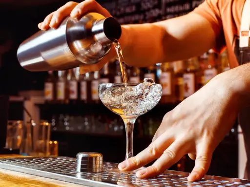 Un barman verse un liquide d'un shaker dans un verre à cocktail rempli de glace. Les shakers sont essentiels pour créer des boissons rafraîchissantes.