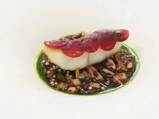 Un poisson blanc délicat, garni de fines lamelles de betterave rouge, repose sur un lit de grains de riz noir et de morceaux de poulpe, le tout nappé d'une sauce verte vibrante. Une œuvre d'art culinaire, un de ces mets d'exception.