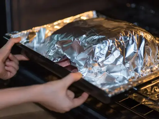 Mains dans le four, une personne place un plat recouvert de papier **alu** pour une cuisson parfaite.