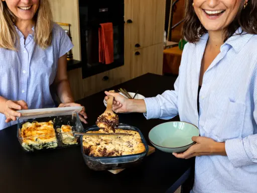 Deux femmes souriantes servent un plat gourmand d'une casserole qui va au four. L'une sert une lasagne, l'autre un dessert chocolaté.