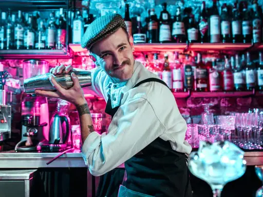 Un barman souriant, casquette vissée, montre comment utiliser un shaker. Derrière lui, des étagères remplies de bouteilles s'illuminent de néons roses et bleus.
