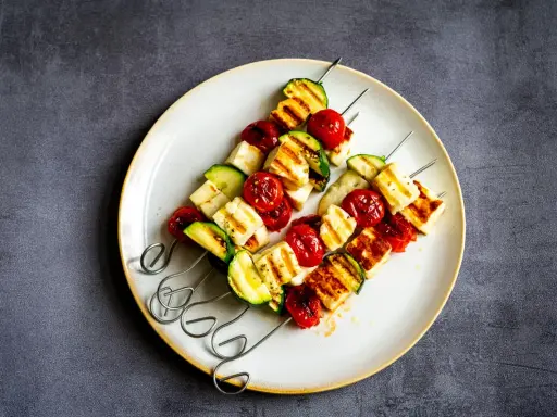 Brochettes de barbecue végétarien : cubes de halloumi grillé, tomates cerises et courgettes sur des brochettes métalliques, servis sur une assiette.