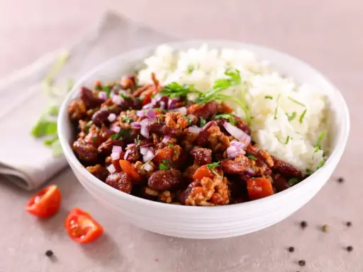Un bol de riz blanc et de chili con carne recette de chef, garni d'oignons rouges et de persil. Des tomates cerises et du poivre en grains décorent la table.