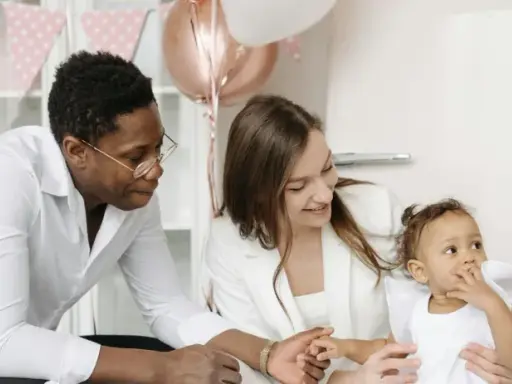 Famille souriante préparant une fête. La mère tient le bébé, le père regarde. Décoration de ballons et guirlandes roses. Une aide pour le check list bapteme.