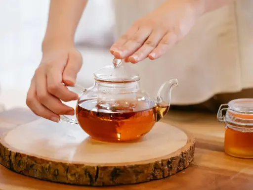 Mains tenant une théière en verre remplie d'un thé ambré, prête à servir le meilleur thé. Un pot de miel à côté.