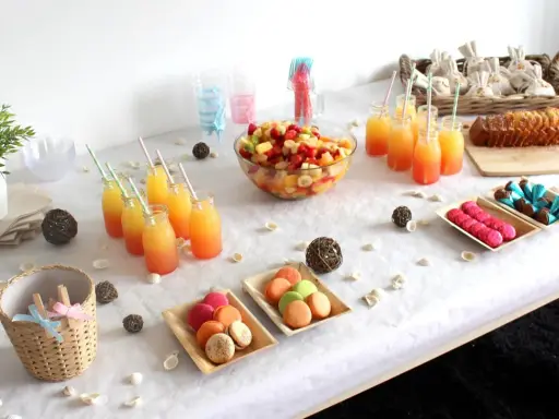 Buffet d'une idée gender reveal : jus de fruits colorés, salade de fruits, macarons roses et verts, gâteau, chocolats bleus et roses.