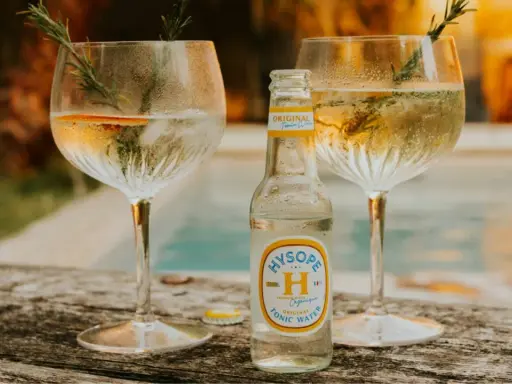 Une bouteille de tonic Hysopé Original, prête à être versée dans deux verres de gin rafraîchissants, décorés de romarin et d'une tranche d'orange.