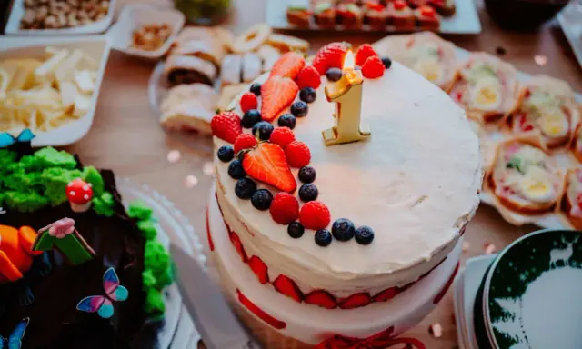 Gâteau à deux étages décoré de fruits rouges et d'une bougie "1" pour un déjeuner d'anniversaire. Un autre gâteau chocolaté, des petits fours et des fruits sont disposés sur la table.