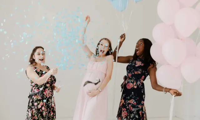 Trois femmes célèbrent un gender reveal, avec des confettis bleus et des ballons roses et bleus. C'est quoi ce moment joyeux !