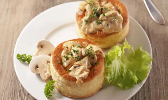 Deux bouchées à la reine maison, garnies d'une sauce crémeuse aux champignons et persil, servies avec des champignons frais et une feuille de laitue.