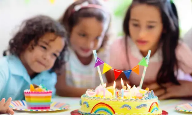 Trois enfants regardent un gâteau d'anniversaire jaune décoré d'arcs-en-ciel et de fanions. Le meilleur goût gâteau anniversaire est prêt à être dégusté.