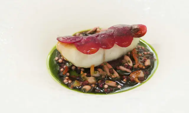 Un poisson blanc délicat, garni de fines lamelles de betterave rouge, repose sur un lit de grains de riz noir et de morceaux de poulpe, le tout nappé d'une sauce verte vibrante. Une œuvre d'art culinaire, un de ces mets d'exception.