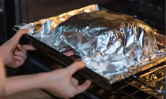 Mains dans le four, une personne place un plat recouvert de papier **alu** pour une cuisson parfaite.