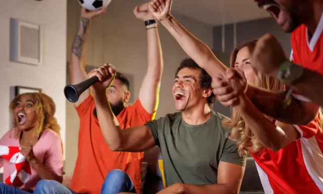 Des supporters en pleine soiree foot, exultant devant leur écran. Un homme lève un ballon, un autre souffle dans une trompette.