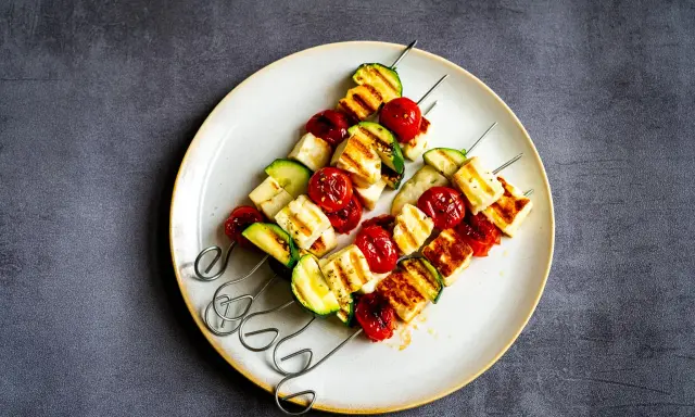 Brochettes de barbecue végétarien : cubes de halloumi grillé, tomates cerises et courgettes sur des brochettes métalliques, servis sur une assiette.