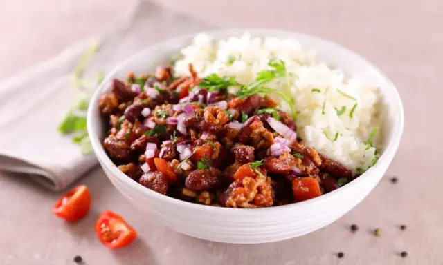Un bol de riz blanc et de chili con carne recette de chef, garni d'oignons rouges et de persil. Des tomates cerises et du poivre en grains décorent la table.