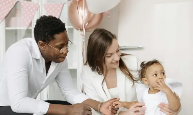 Famille souriante préparant une fête. La mère tient le bébé, le père regarde. Décoration de ballons et guirlandes roses. Une aide pour le check list bapteme.