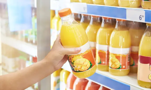 Une main choisit une bouteille de ce qui semble être le meilleur jus de fruit, avec des mangues et des ananas sur l'étiquette.