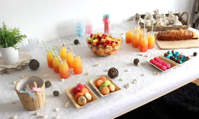 Buffet d'une idée gender reveal : jus de fruits colorés, salade de fruits, macarons roses et verts, gâteau, chocolats bleus et roses.