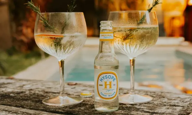 Une bouteille de tonic Hysopé Original, prête à être versée dans deux verres de gin rafraîchissants, décorés de romarin et d'une tranche d'orange.