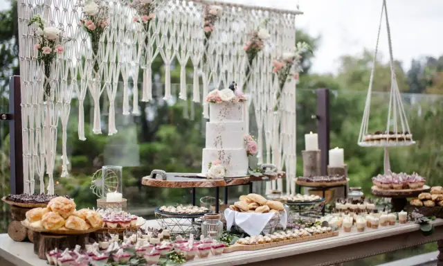 Un buffet de mariage bohème avec un gâteau à trois étages, des petits fours, des verrines et des pains. Une décoration macramé et florale complète cette idée repas mariage.