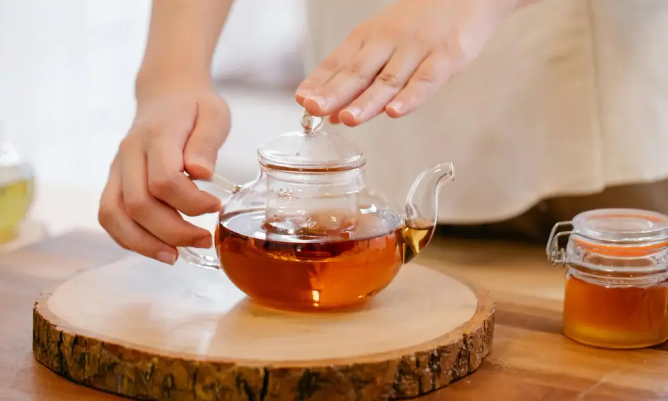 Mains tenant une théière en verre remplie d'un thé ambré, prête à servir le meilleur thé. Un pot de miel à côté.
