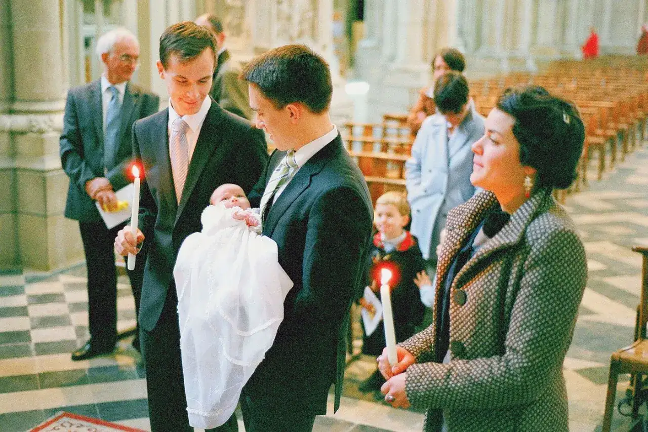 Un bébé en tenue blanche est tenu par son parrain, une bougie allumée à la main. La marraine est à ses côtés. C'est une scène d'animation baptême parrain marraine.