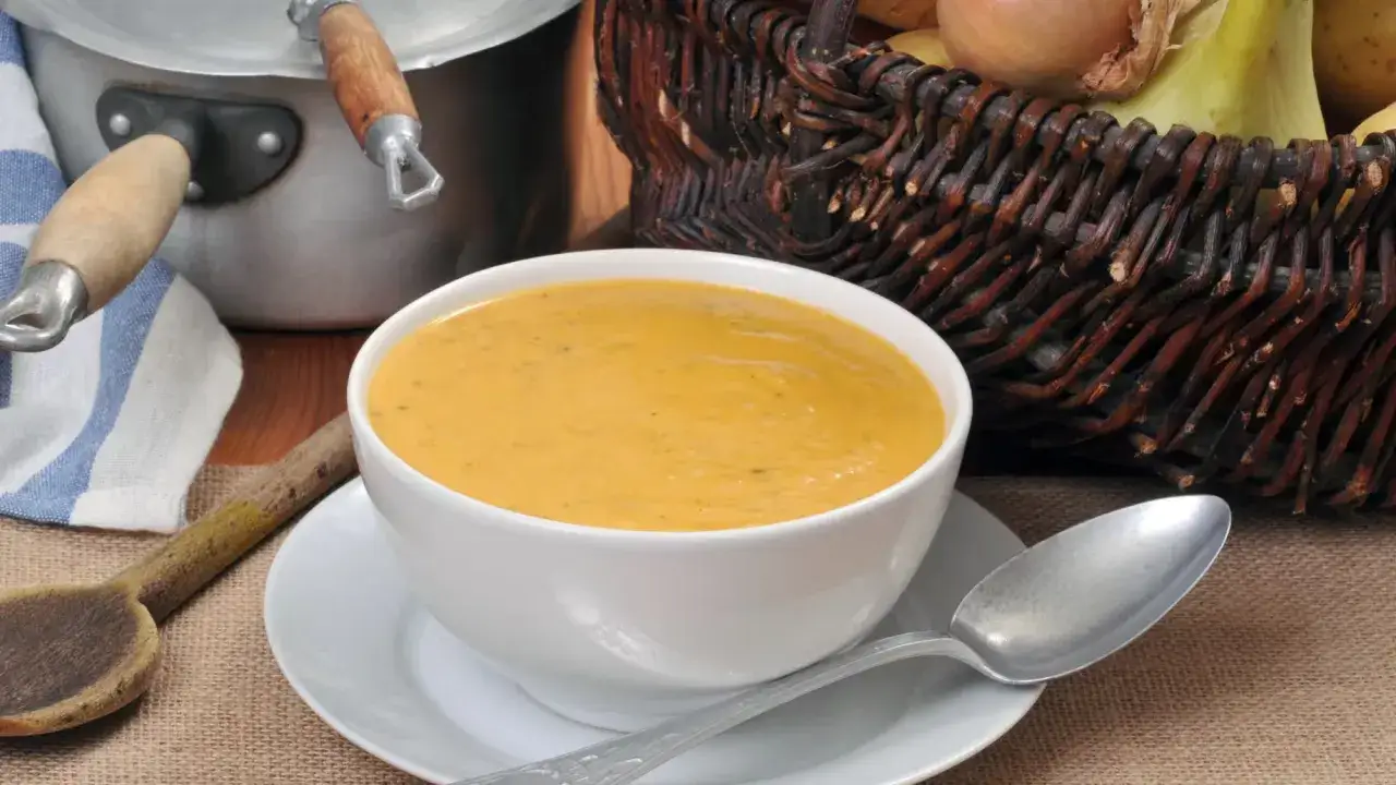 Un bol de soupe de légumes variés, onctueuse et réconfortante, est prêt à être dégusté. Une cuillère en bois et un panier de légumes complètent la scène.