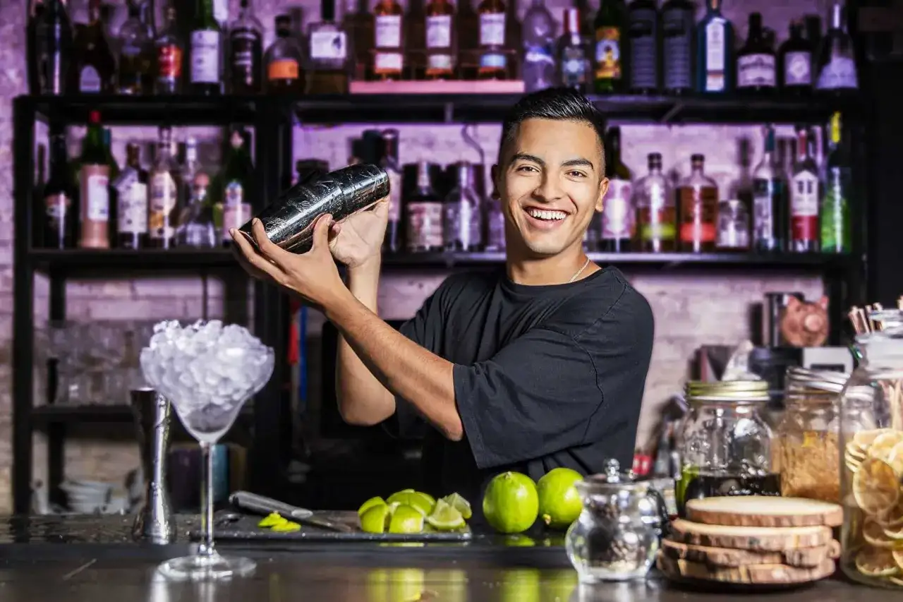 Un barman souriant montre comment utiliser un shaker pour préparer un cocktail. Des limes et de la glace sont prêts.