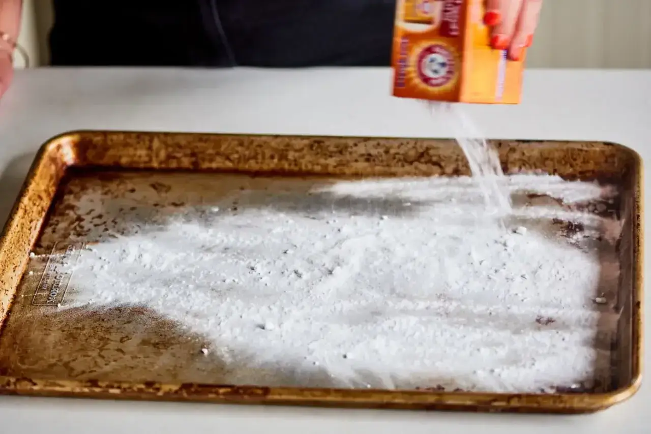 Une main verse du bicarbonate de soude sur une plaque de cuisson sale pour montrer comment dégraisser un plat.