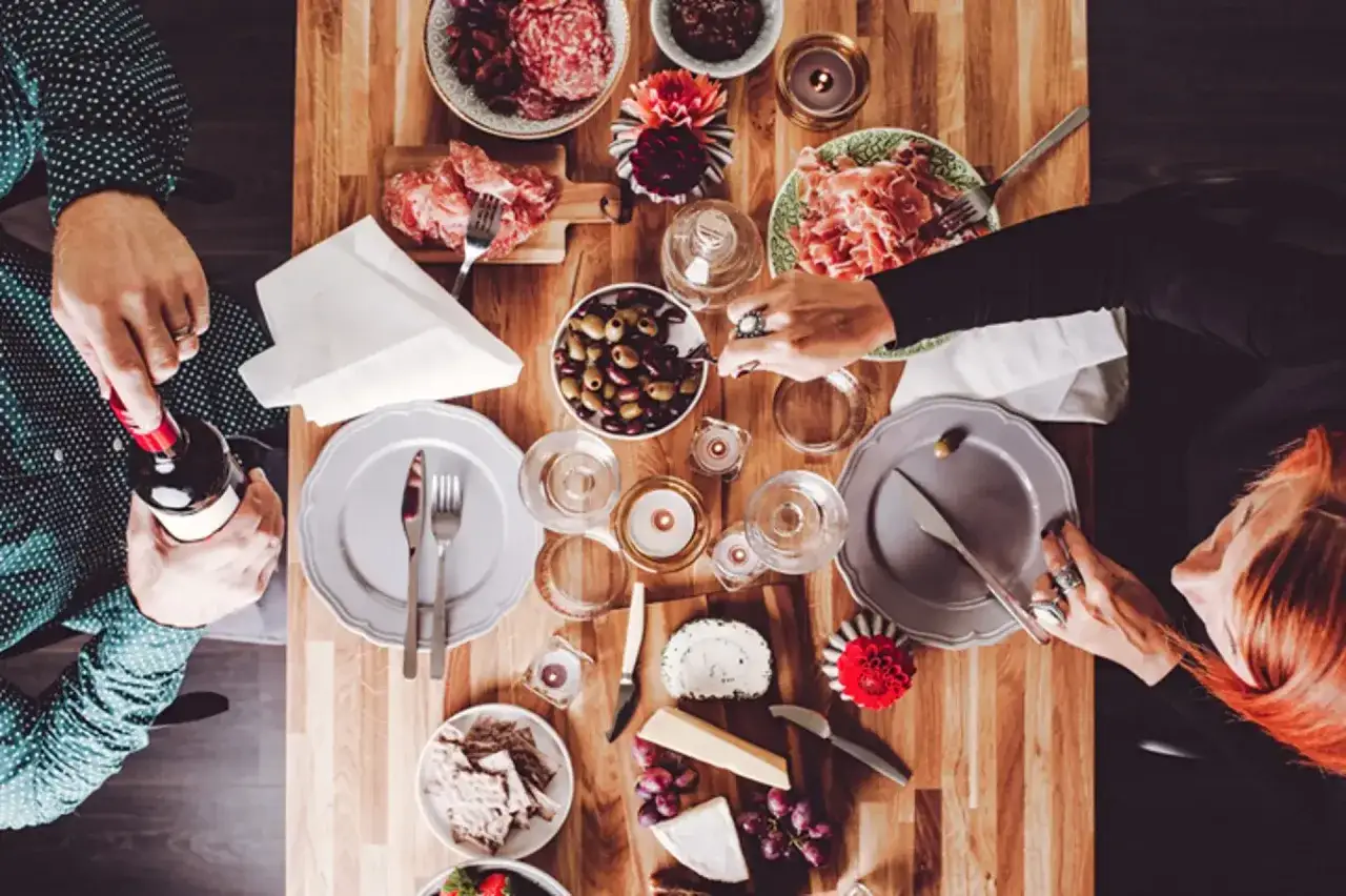 Un homme ouvre une bouteille de vin pour un déjeuner convivial. Sur la table en bois, charcuterie, olives, fromages, raisins et crackers invitent au partage.