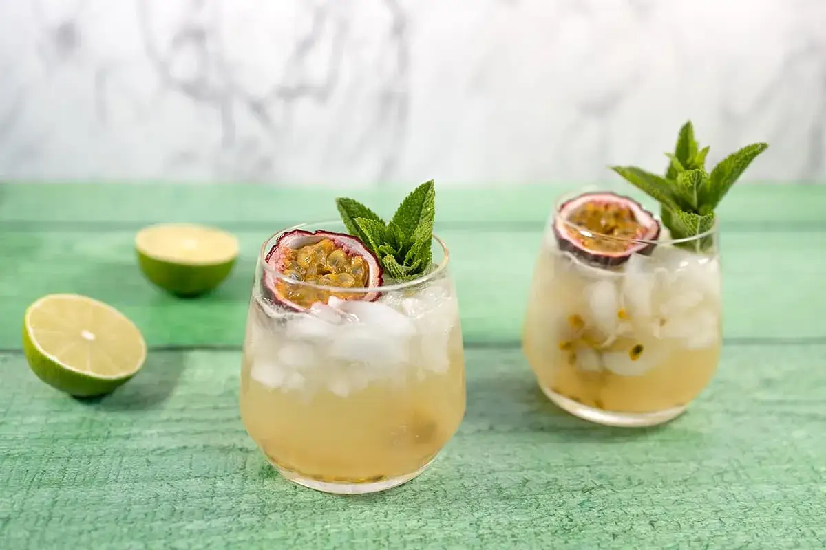 Deux verres de cocktail sans alcool à préparer la veille, garnis de fruits de la passion et de menthe, avec des citrons verts coupés en arrière-plan.