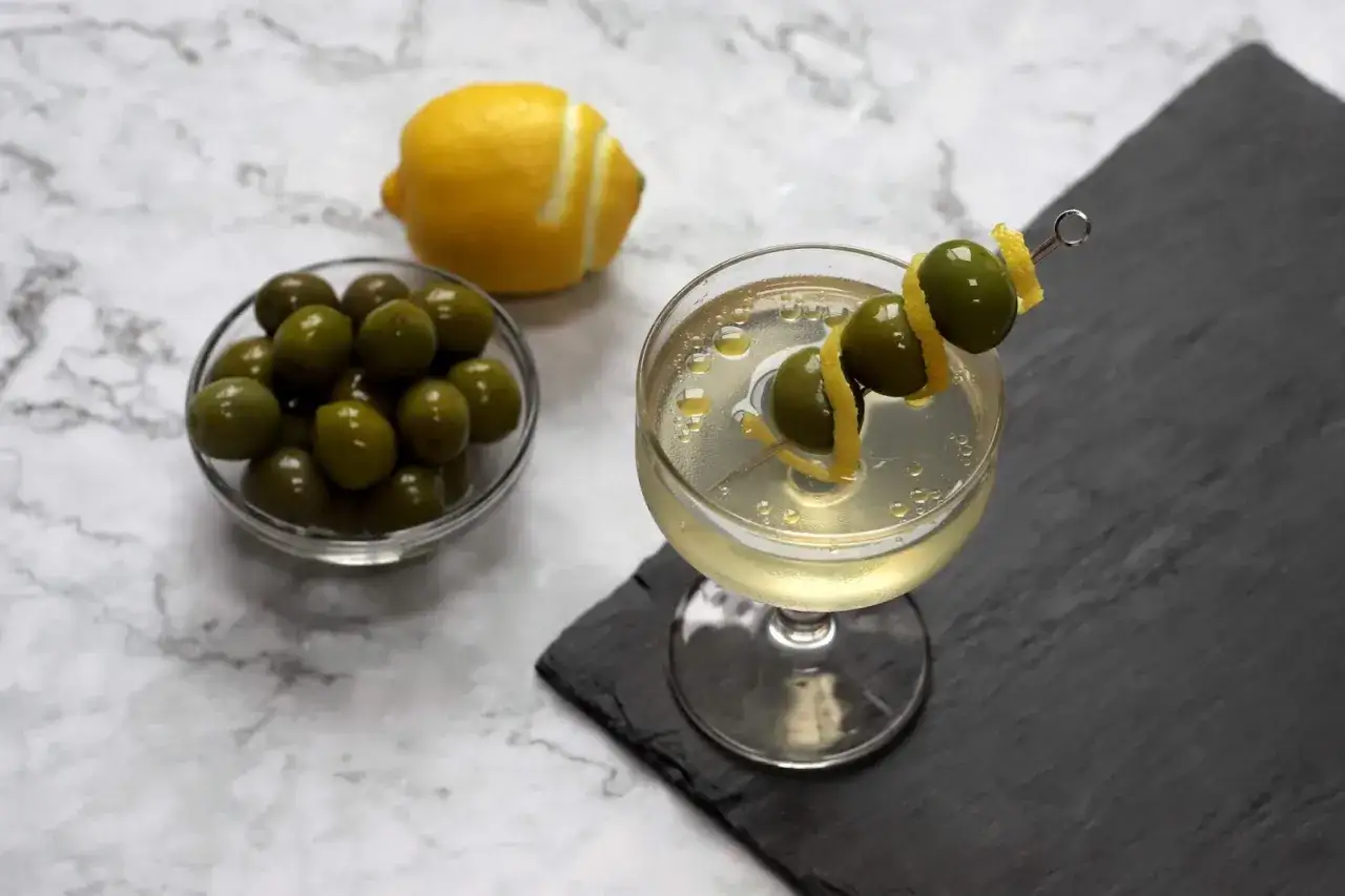Un verre de martini avec trois olives sur une brochette et un zeste de citron. &Agrave; c&ocirc;t&eacute;, un bol d'olives et un citron.