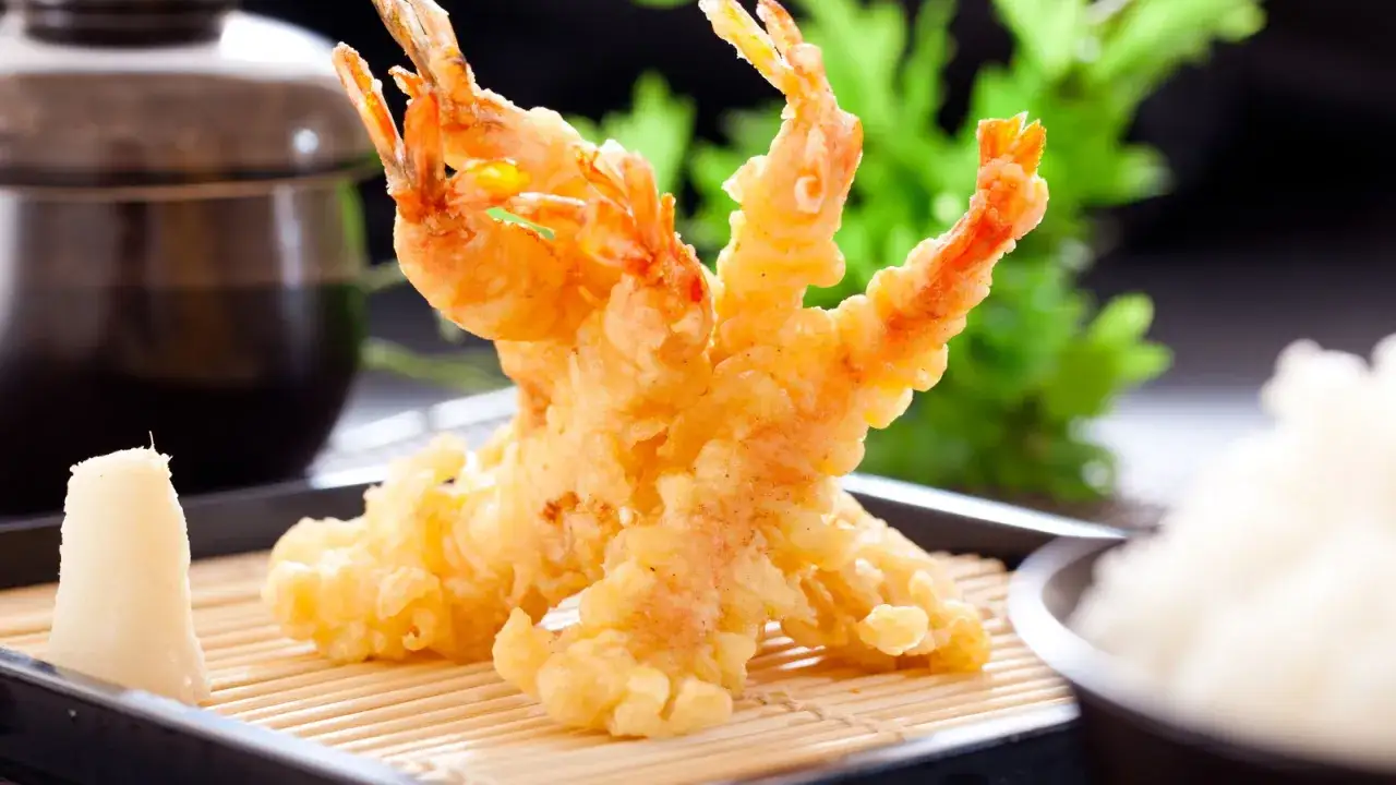 Deux crevettes en tempura dorées et croustillantes, servies sur un lit de bambou avec du riz blanc et une sauce foncée.