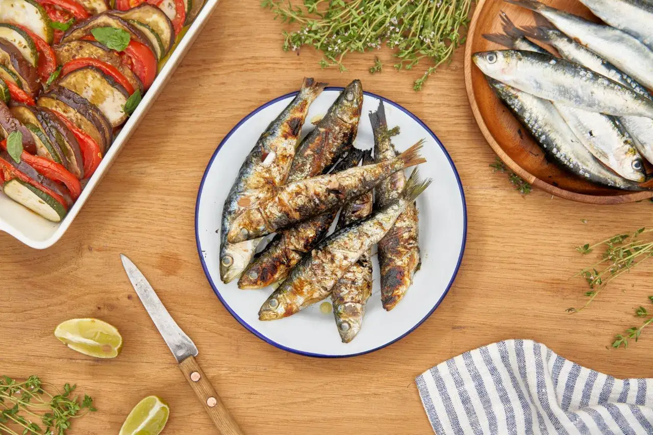 Sardines grillées sur une assiette, accompagnées d'un plat de légumes et de citrons. Une belle idée pour une sardinade recette.