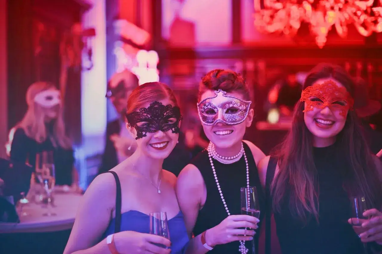 Trois femmes masquées sourient, verre à la main, lors d'une soirée costumée. L'ambiance est festive et mystérieuse.
