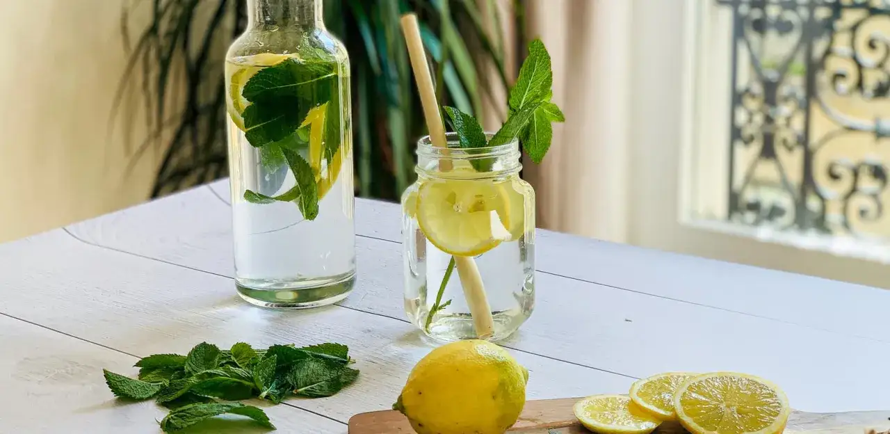 Bouteille et verre remplis de citronnade maison rafraîchissante avec des tranches de citron et des feuilles de menthe. Des citrons et de la menthe fraîche sont disposés sur la table.