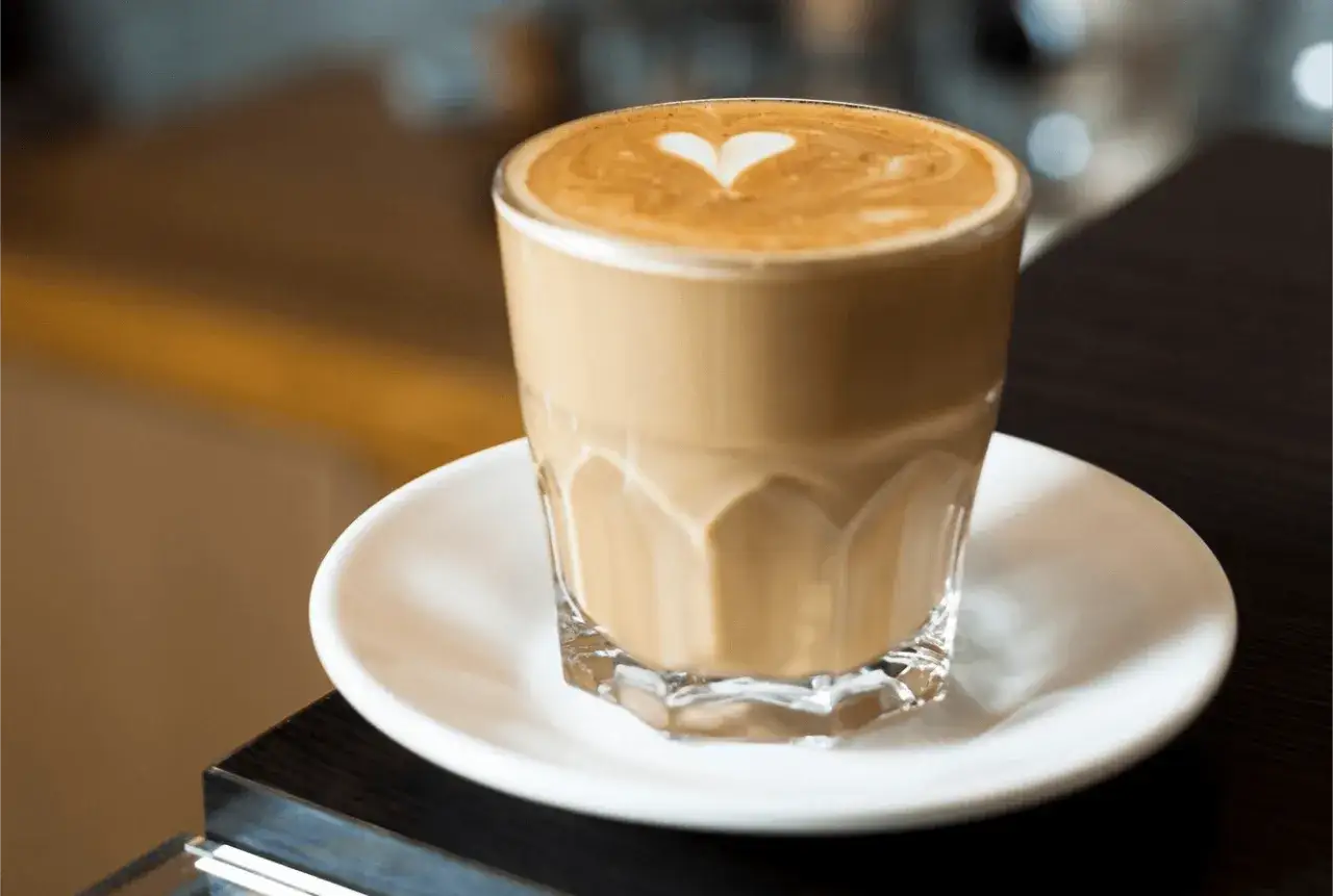 Un café latte crémeux avec un cœur dessiné sur la mousse, servi dans un verre transparent sur une soucoupe blanche.