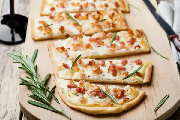 Quatre tartes flambées, une délicieuse entrée alsacienne, garnies de lardons et de romarin sur une planche en bois.